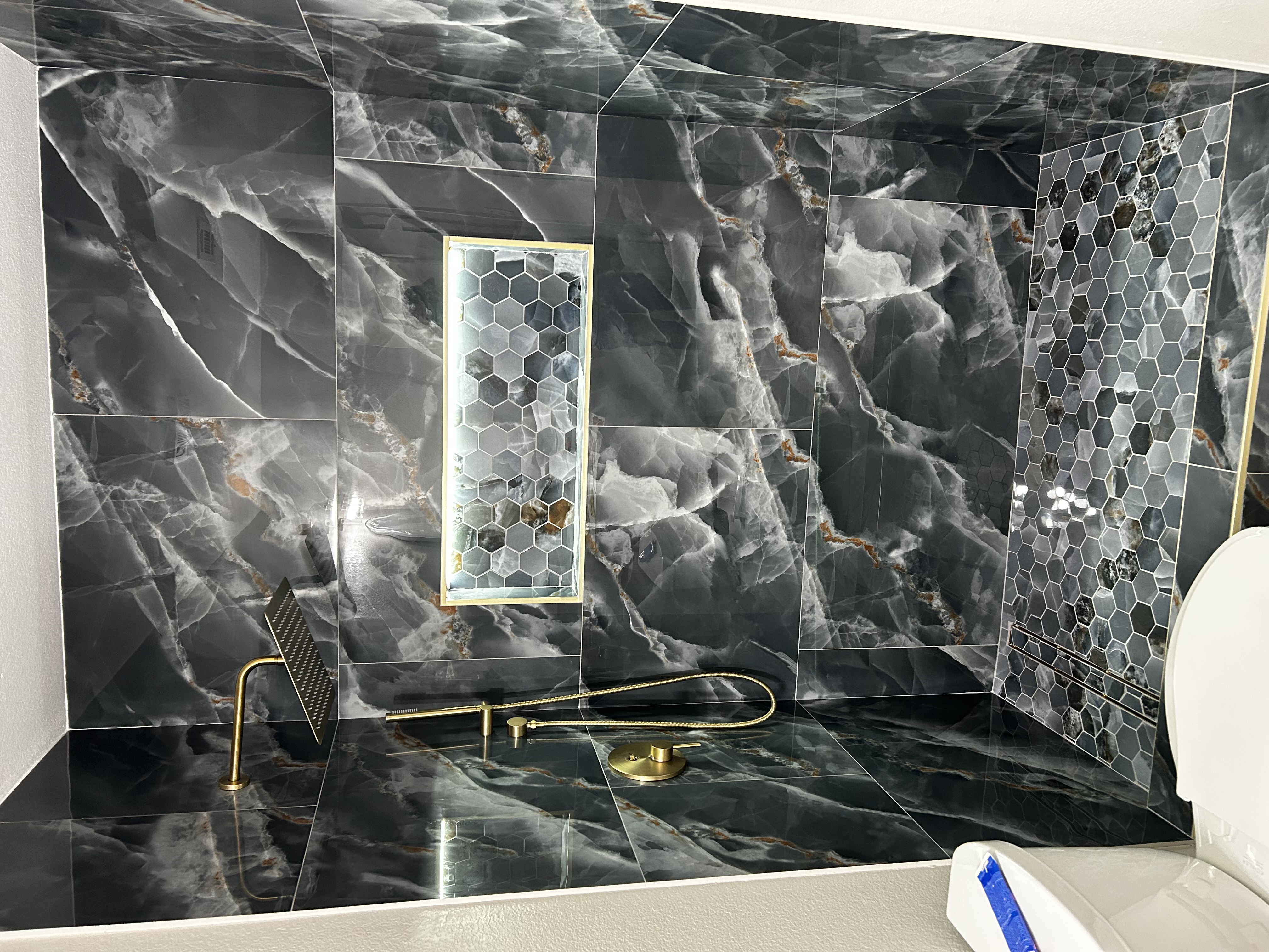 Luxury bathroom remodel by HN Construction with dark onyx-look marble tile, a lit hexagon shower niche, and brushed-gold fixtures
